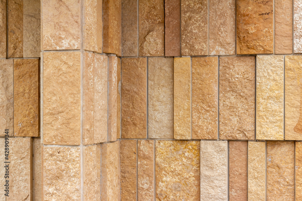 Corner angle view of a modern style rough textured limestone brick wall background with attractive vertical aligned natural kasota stone blocks in varying widths and shades of light brown. Corner angle view of a modern style rough textured limestone brick wall background with attractive vertical aligned natural kasota stone blocks in varying widths and shades of light brown.