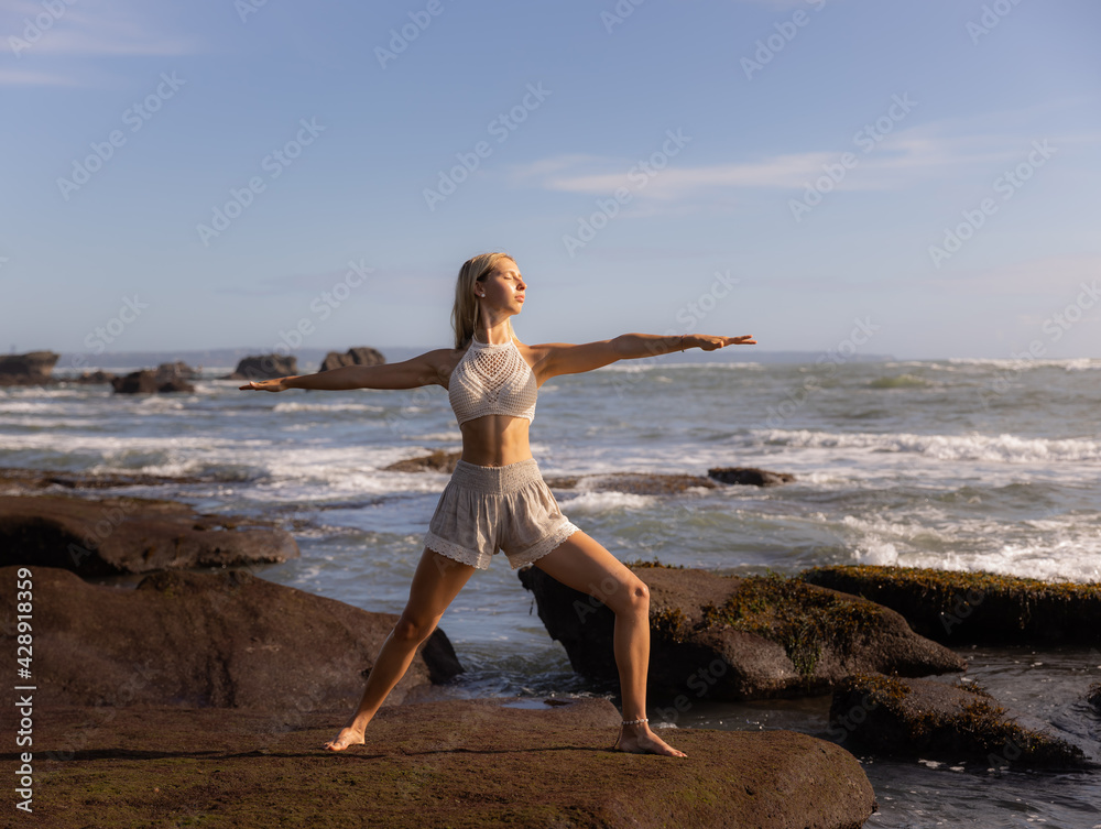 Young woman practicing yoga. Standing in Virabhadrasana II pose, Warrior II Pose. Work out. Healthy lifestyle. Strong body. Ocean background. Yoga retreat. Self-care concept. Beach in Bali