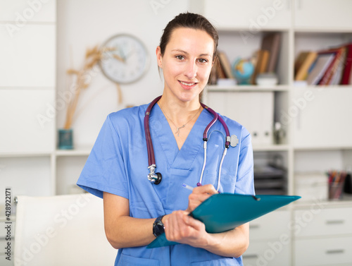 Positive young female doctor working in a clinic takes important notes while standing in a well-lit office © JackF