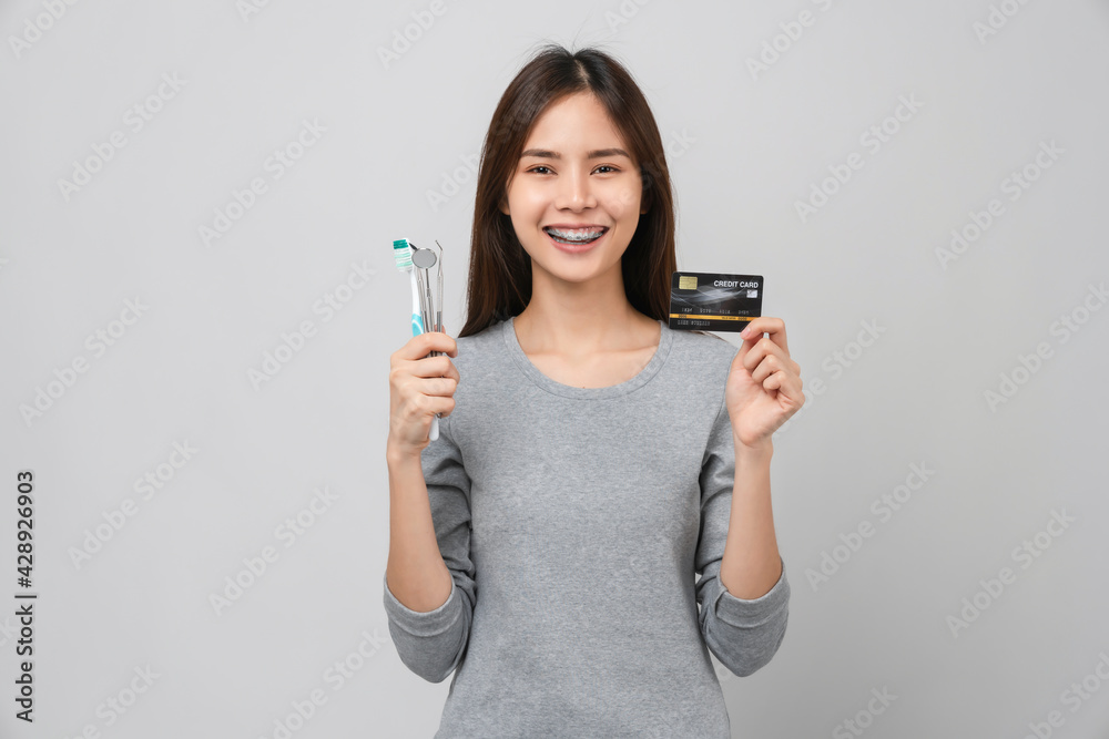 Smiling Asian woman wearing braces with tooth and holding toothbrush and dentist tool, credit card on grey background, Concept oral hygiene and health care. Smiling Asian woman wearing braces with tooth and holding toothbrush and dentist tool, credit card on grey background, Concept oral hygiene and health care.