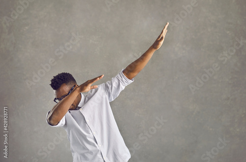 Happy handsome young black man with Afro hairstyle dancing dub against gray stone street wall. Funky African American guy in white shirt doing popular dab dance arm move on concrete urban background photo