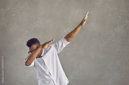 Happy handsome young black man with Afro hairstyle dancing dub against gray stone street wall. Funky African American guy in white shirt doing popular dab dance arm move on concrete urban background photo
