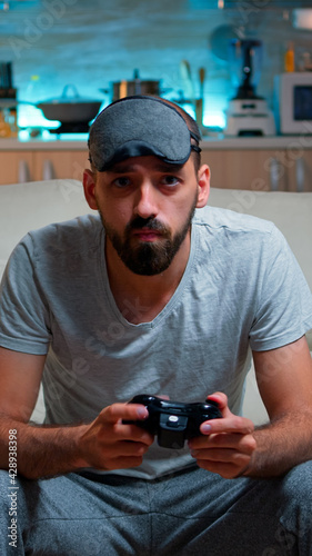 Front view of pro gamer sitting on sofa in front of television while playing videogames competition using wireless joystick. Cheerful man with sleep mask enjoying time alone late at night in kitchen photo