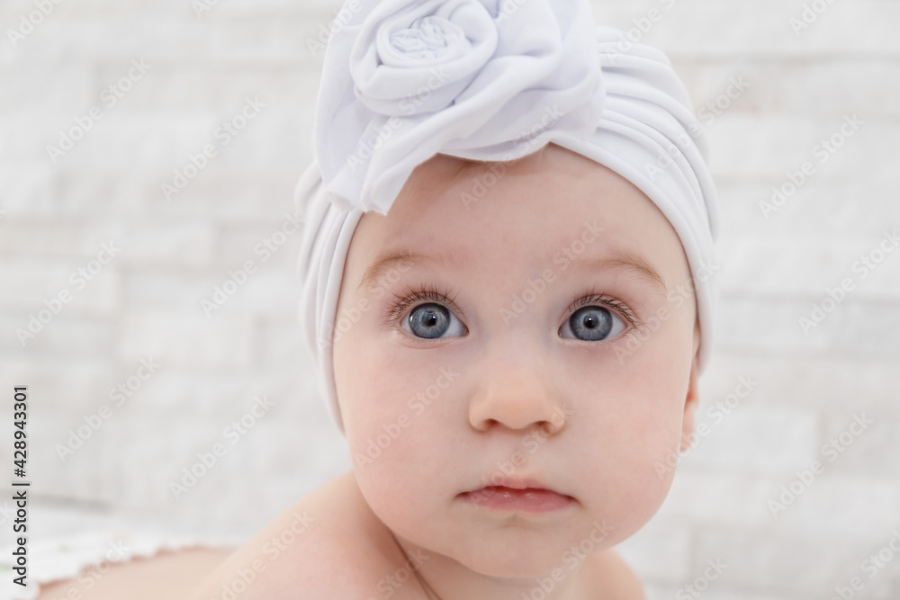 Close-up of cute little funny caucasian baby girl wearing a white turban hat at home, light interior Close-up of cute little funny caucasian baby girl wearing a white turban hat at home, light interior
