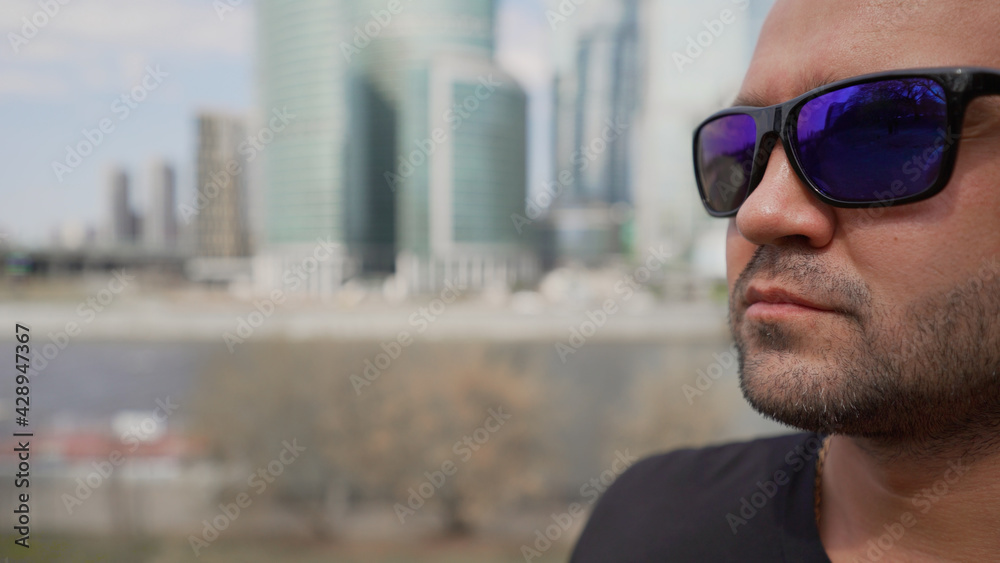 Portrait of brutal man in sunglasses in the city on the background of large building. Brutal guy in sunglasses on the background of the city. Brutal guy on the background of buildings in Moscow.