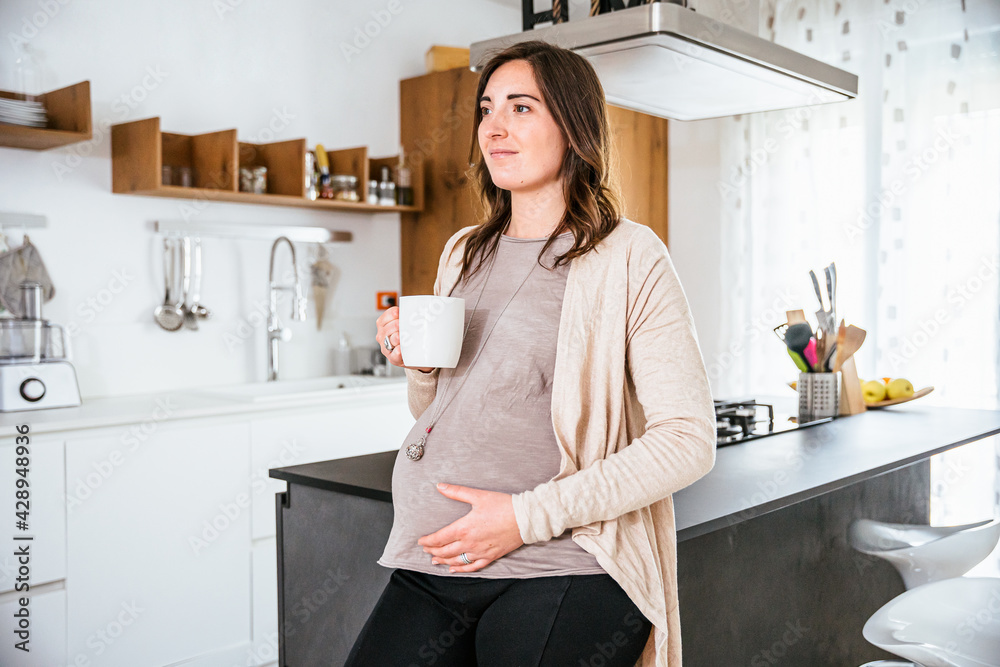 Young pregnant woman in the modern kitchen at home drinks an herbal tea leaning against the cabinet and touches her belly smiling - happy millennial will soon become a mother - Motherhood concept