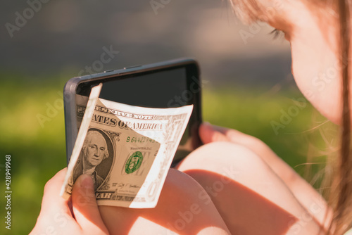 Young beautiful girl holding smartphone andUS Dollar bill as symbol of online payment and shopping. Summer vacation, holiday and quarantine are not an obstacle to normal lifestyle. photo