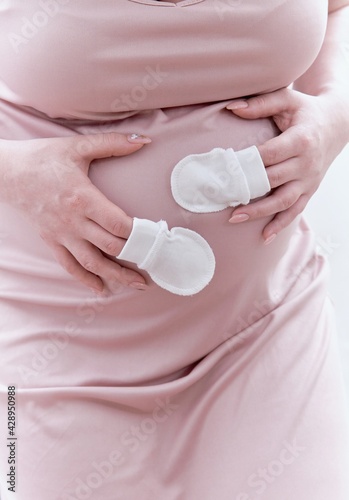 Small shoes for an unborn baby in the belly of a pregnant woman. Pregnant woman holding small baby shoes resting at home in the bedroom.