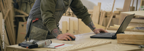 Concentrated adult craftsman checking information on laptop while working in carpentry workshop. profession, carpentry, technology and people concept photo