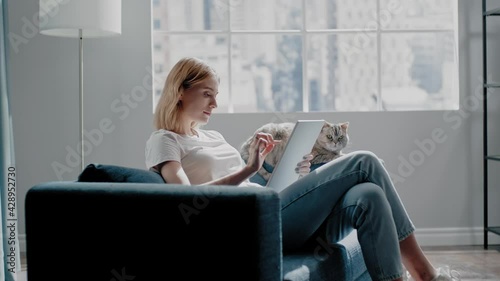 Lady works on tablet near fluffy cat sitting on blue couch