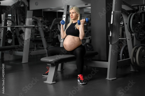 Pregnant woman lifting up dumbbells training biceps muscle at the gym sitting bench