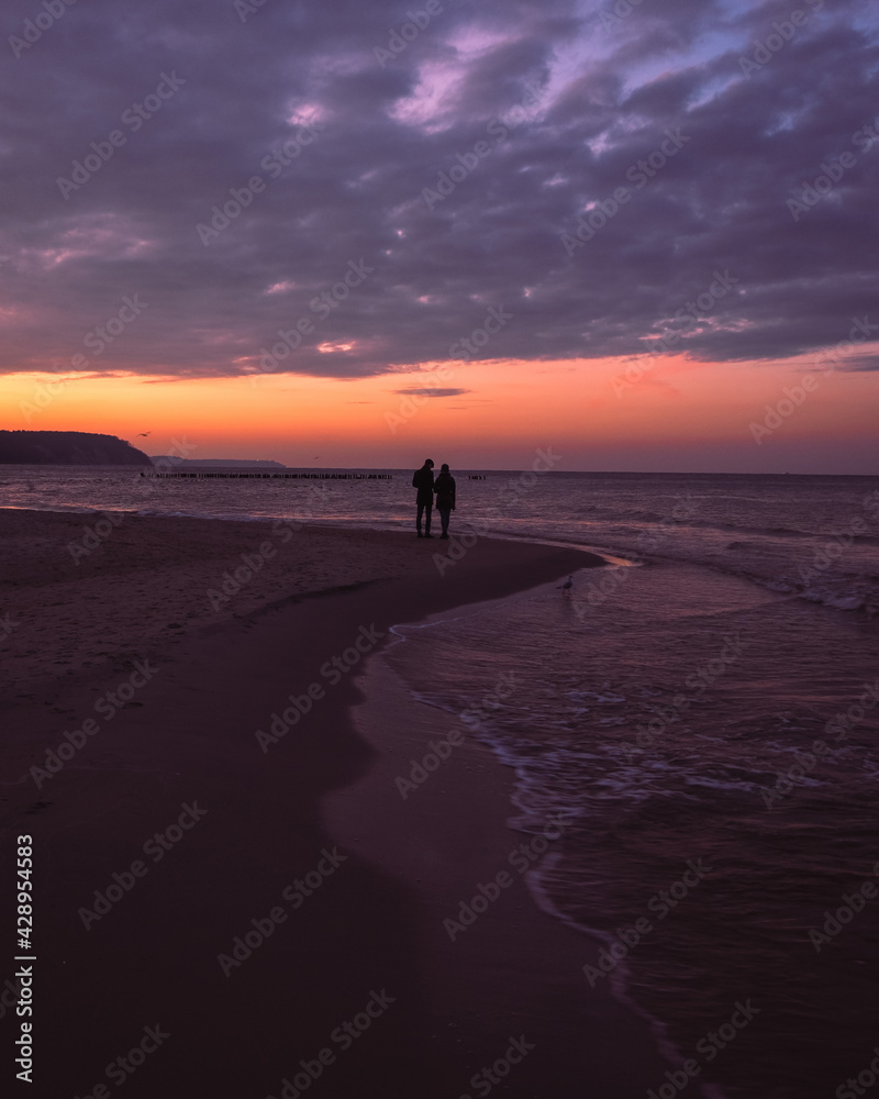 Sunset at the sea. Silhouette of romantic couple standing at looking at breathtaking seascape. Beautiful view on the beach and water, purple sky with clouds, vacation at Baltic sea. Ocean in the dusk.