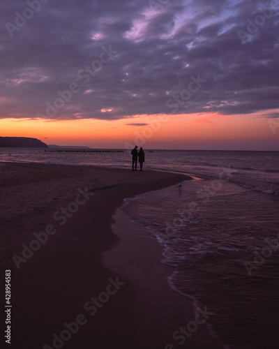 Sunset at the sea. Silhouette of romantic couple standing at looking at breathtaking seascape. Beautiful view on the beach and water, purple sky with clouds, vacation at Baltic sea. Ocean in the dusk.