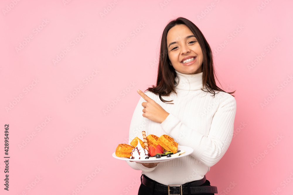 Young brunette woman holding waffles over isolated pink background pointing to the side to present a product Young brunette woman holding waffles over isolated pink background pointing to the side to present a product