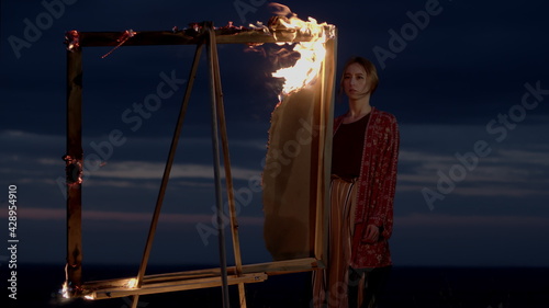 Young Girl Artist Stands In Nature At Dusk In Front Of An Easel With Burning Picture.