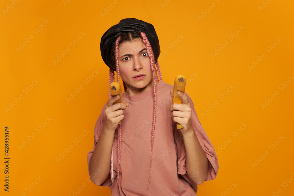 Studio shot of fashionable cute young Caucasian woman wearing synthetic box braids staring at camera with angry look frowning eyebrows, aiming two bananas at you as if shooting guns, playing Studio shot of fashionable cute young Caucasian woman wearing synthetic box braids staring at camera with angry look frowning eyebrows, aiming two bananas at you as if shooting guns, playing