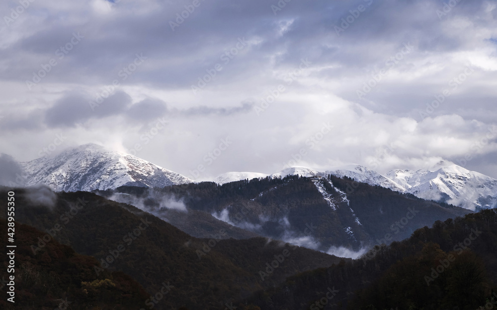 Snow peaks of the mountains and cloudy sky on the background. Beautiful scenery, outdoor snowy view. Green hills.