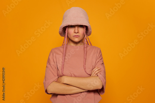 Trendy looking young female with ring in her nose posing over yellow wall wearing stylish teddy bear hat and oversized t-shirt crossing arms on her chest having confident serious facial expression