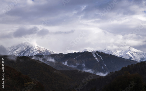 Snow peaks of the mountains and cloudy sky on the background. Beautiful scenery, outdoor snowy view. Green hills. © Derariad