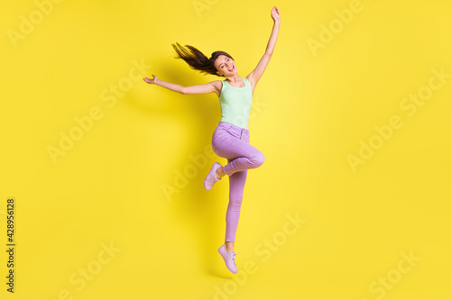 Photo of charming positive lady raise hands jump wear green top pants footwear isolated yellow color background