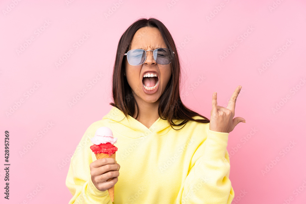 Young brunette girl holding a cornet ice cream over isolated pink background making rock gesture Young brunette girl holding a cornet ice cream over isolated pink background making rock gesture