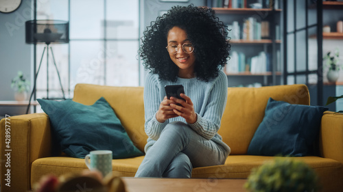 Portrait of a Beautiful Authentic Latina Female in a Stylish Cozy Living Room Using Smartphone at Home. She's Browsing the Internet and Checking Videos on Social Networks and Having Fun. photo