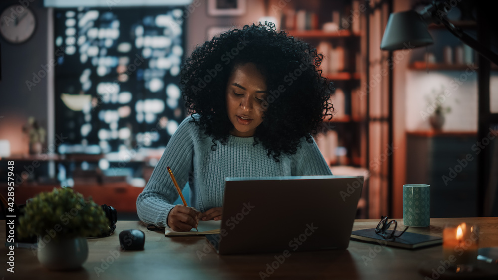 Beautiful Authentic Latina Female Sitting at a Desk in a Cozy Dark Living Room in the Evening and Using Laptop Computer at Home. Student Studying in Home School. Chat with Friends on Social Network. Beautiful Authentic Latina Female Sitting at a Desk in a Cozy Dark Living Room in the Evening and Using Laptop Computer at Home. Student Studying in Home School. Chat with Friends on Social Network.