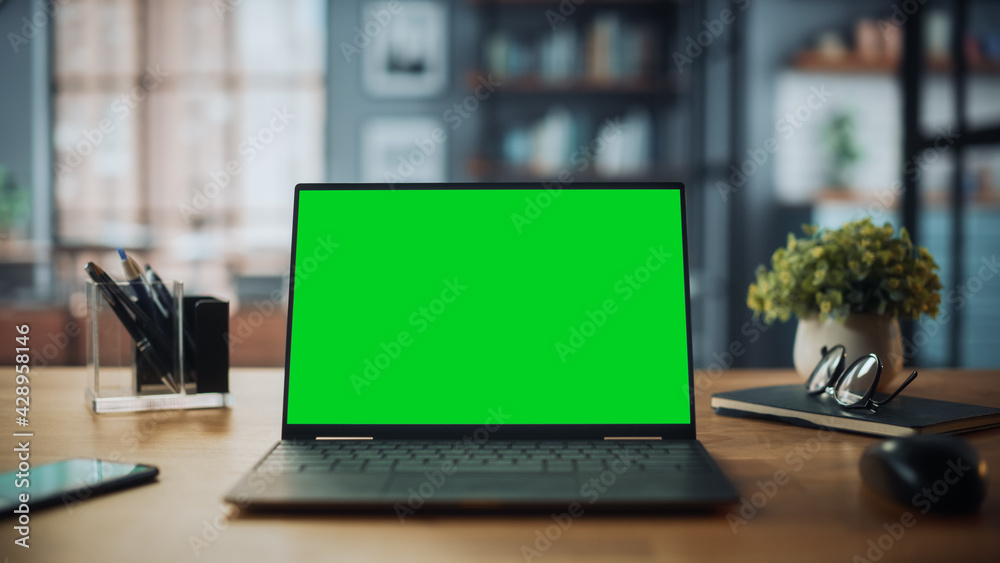 Laptop Computer with Mock Up Green Screen Display Standing on the Table in Cozy Living Room. In the Background Stylish Modern Bright Home Office Studio During Day with Big Cityscape Window. Laptop Computer with Mock Up Green Screen Display Standing on the Table in Cozy Living Room. In the Background Stylish Modern Bright Home Office Studio During Day with Big Cityscape Window.