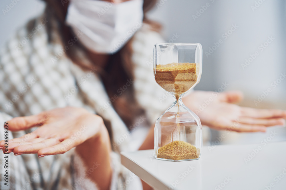 Adult woman in mask feeling sick at home with hourglass Covid-19 concept Adult woman in mask feeling sick at home with hourglass Covid-19 concept