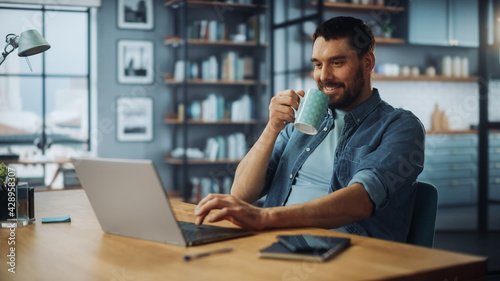 Handsome Caucasian Man Working on Laptop Computer while Sitting on a Sofa Couch in Stylish Cozy Living Room. Freelancer Working From Home. Browsing Internet, Drinking Coffee from a Mug, Having Fun.