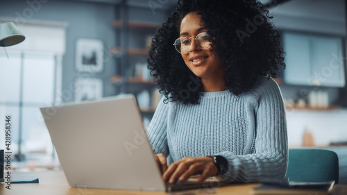 Beautiful Authentic Latina Female with Afro Hair Sitting at a Desk in a Cozy Living Room and Using Laptop Computer at Home. She's Browsing the Internet and Checking Videos on Social Networks.