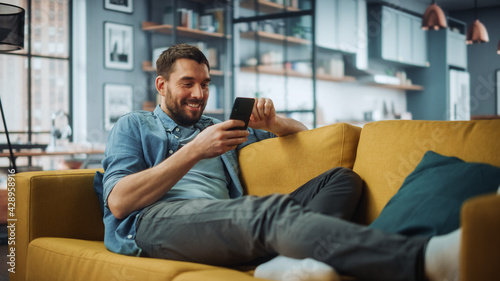 Happy Handsome Caucasian Man Using Smartphone in Cozy Living Room at Home. Man Resting on Comfortable Sofa. He's Browsing the Internet and Checking Videos on Social Networks and Having Fun. photo