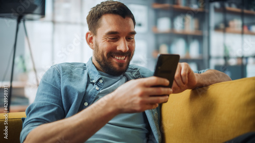 Happy Handsome Caucasian Man Using Smartphone in Cozy Living Room at Home. Man Resting on Comfortable Sofa. He's Browsing the Internet and Checking Videos on Social Networks and Having Fun.