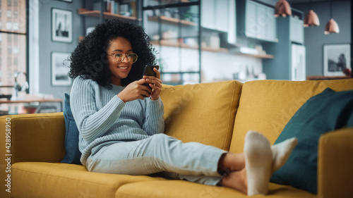 Happy Beautiful Latina Female Using Smartphone in Cozy Living Room at Home. Female Resting on Comfortable Sofa. She's Browsing the Internet and Checking Videos on Social Networks and Having Fun.