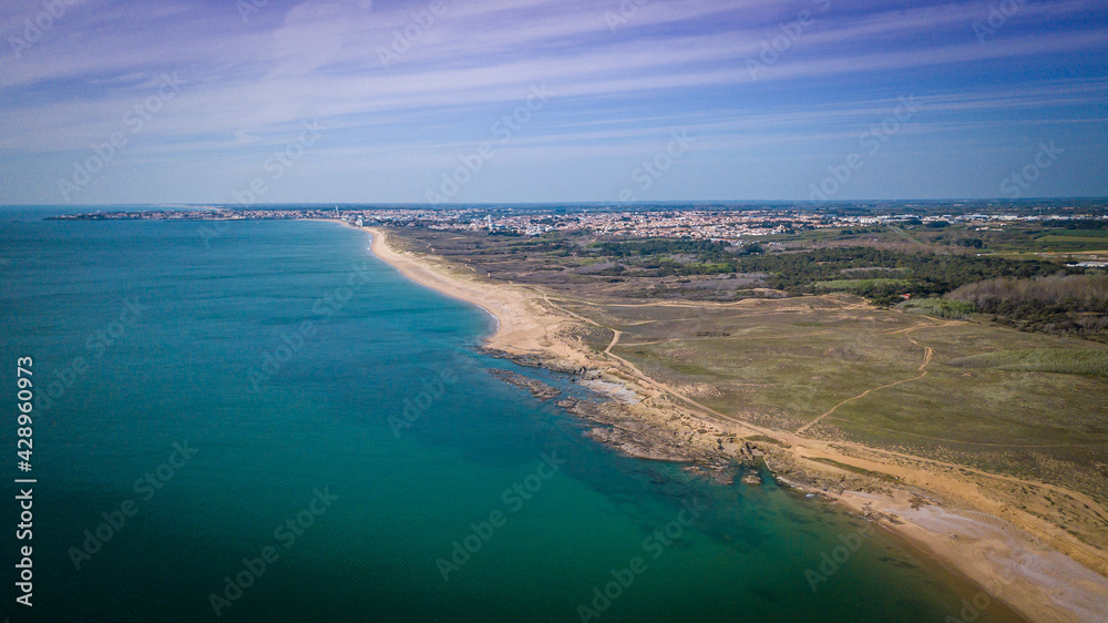 Aerial photography of sunny beaches from above with clear water, sandy dunes, rocks and a town in the background with its harbor
