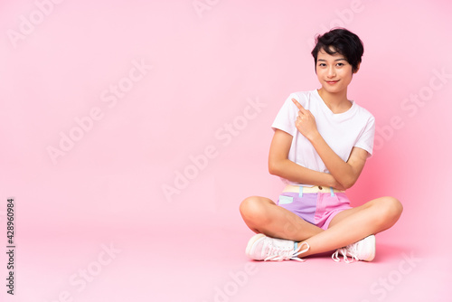 Young Vietnamese woman with short hair sitting on the floor over isolated pink background pointing to the side to present a product