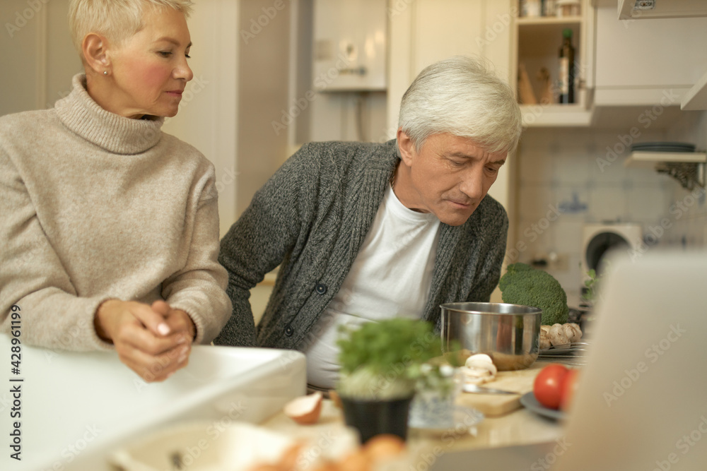 Food, nutrition, diet, people and retirement concept. Serious gray haired male pensioner and his wife experimenting in kitchen, making healthy lunch using cooking utensils and fresh products Food, nutrition, diet, people and retirement concept. Serious gray haired male pensioner and his wife experimenting in kitchen, making healthy lunch using cooking utensils and fresh products