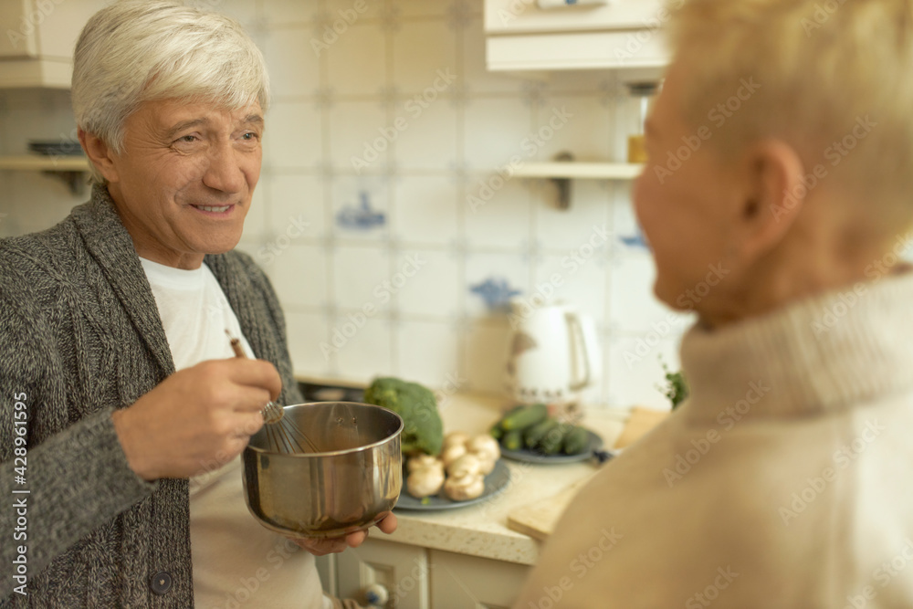 Attractive Caucasian man with gray hair holding saucepan and whisk stirring ingredients for dough making vegetarian pizza. Handsome retired male talking to his wife while cooking dinner in kitchen