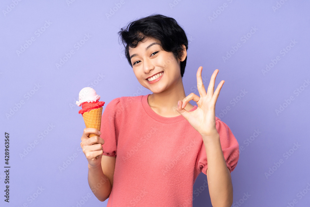 Young Vietnamese woman with short hair holding a cornet ice cream over isolated purple background showing ok sign with fingers Young Vietnamese woman with short hair holding a cornet ice cream over isolated purple background showing ok sign with fingers