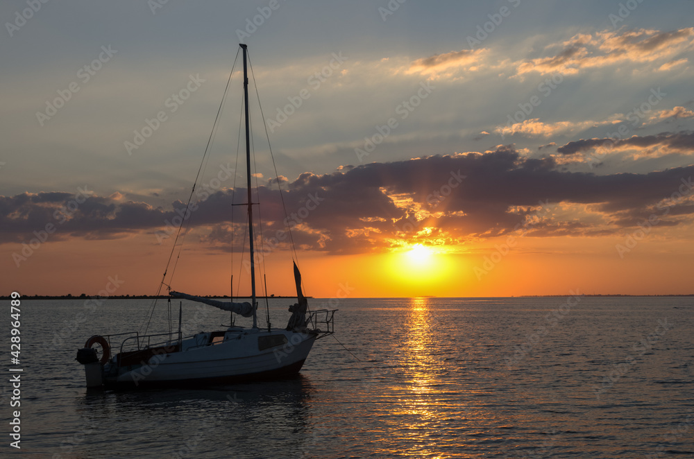 Small sailboat docked by the coast at sunset, Black sea. Family vacation or travel by sea on own yacht. Beautiful evening sky with beams of sun through clouds. Big sun over water in the evening.