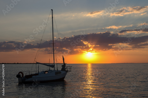 Small sailboat docked by the coast at sunset, Black sea. Family vacation or travel by sea on own yacht. Beautiful evening sky with beams of sun through clouds. Big sun over water in the evening. © Kseniia