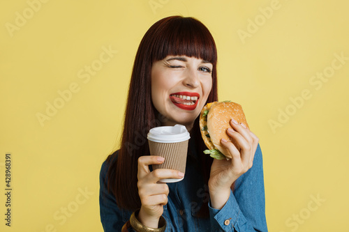 Close up of a cheerful young attractive woman grimacing at camera  showing tongue  holding tasty burger and take away coffee and enjoying her lunch time. Isolated on yellow studio backgroud