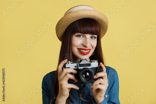 Stylish red haired smiling woman photographer , wearing straw hat and denim shirt, posing with retro camera on the yellow wall background. Image with copy space.