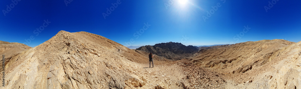male hiker on a hiking trail in Eilat mountains. Colorful nature pattern painted on a rock wall. Rock formations and boulders. Panoramic view over the trail on surrounding red mountains