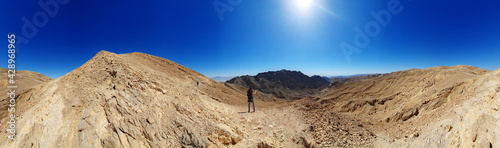 male hiker on a hiking trail in Eilat mountains. Colorful nature pattern painted on a rock wall. Rock formations and boulders. Panoramic view over the trail on surrounding red mountains