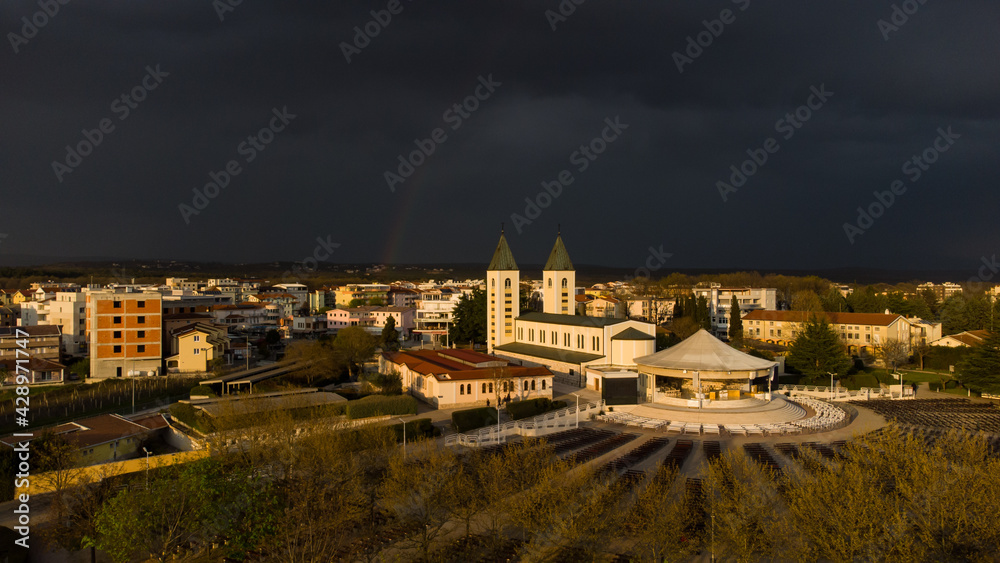 Aerial drone view of Medjugorje. Catholic Church illuminated by the last sun rays at sunset. Dark clouds of a storm and rainbow in distance. Saint James church in Međugorje, Bosnia and Herzegovina. 
