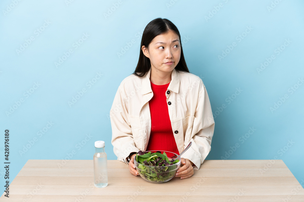 Young Chinese girl eating a salad making doubts gesture looking side Young Chinese girl eating a salad making doubts gesture looking side