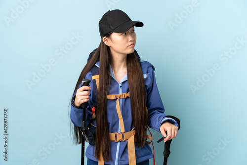 Young Chinese girl with backpack and trekking poles over isolated blue background looking to the side