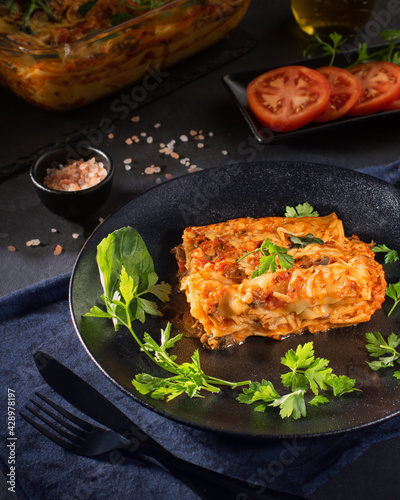 Piece of tasty hot lasagna served with a green leaves on a black plate. Italian cuisine, menu, recipe. Close up, side view.
