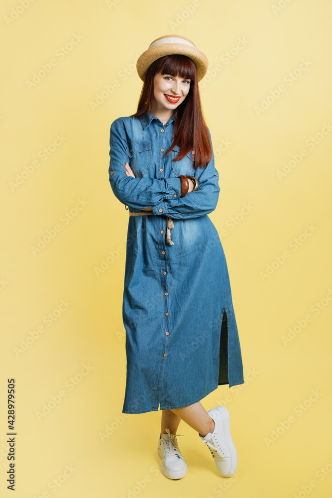 Confident smiling girl posing at camera, isolated on yellow background. Studio shot of fashionable woman in jeans dress and straw hat, posing with crossed arms. Full length portrait. Confident smiling girl posing at camera, isolated on yellow background. Studio shot of fashionable woman in jeans dress and straw hat, posing with crossed arms. Full length portrait.